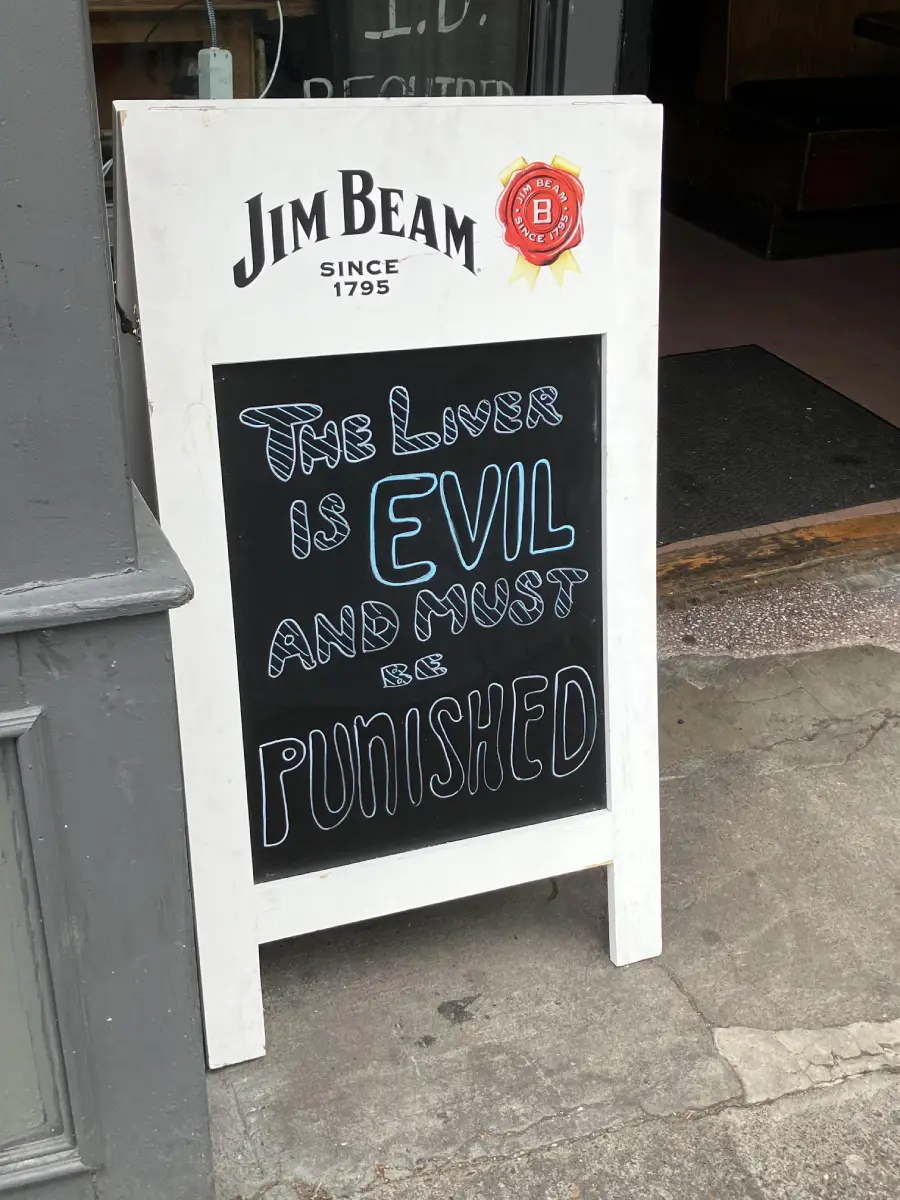 A sidewalk chalkboard sign reads: “The liver is evil and must be punished,” under a Jim Beam logo outside a bar entrance.