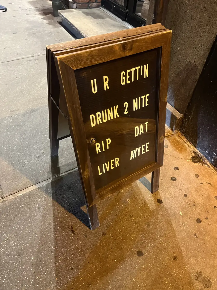 A sidewalk sign reads: “U R GETTIN’ DRUNK 2 NITE RIP DAT LIVER AYYEE” in white letters on a black background with a wooden frame.