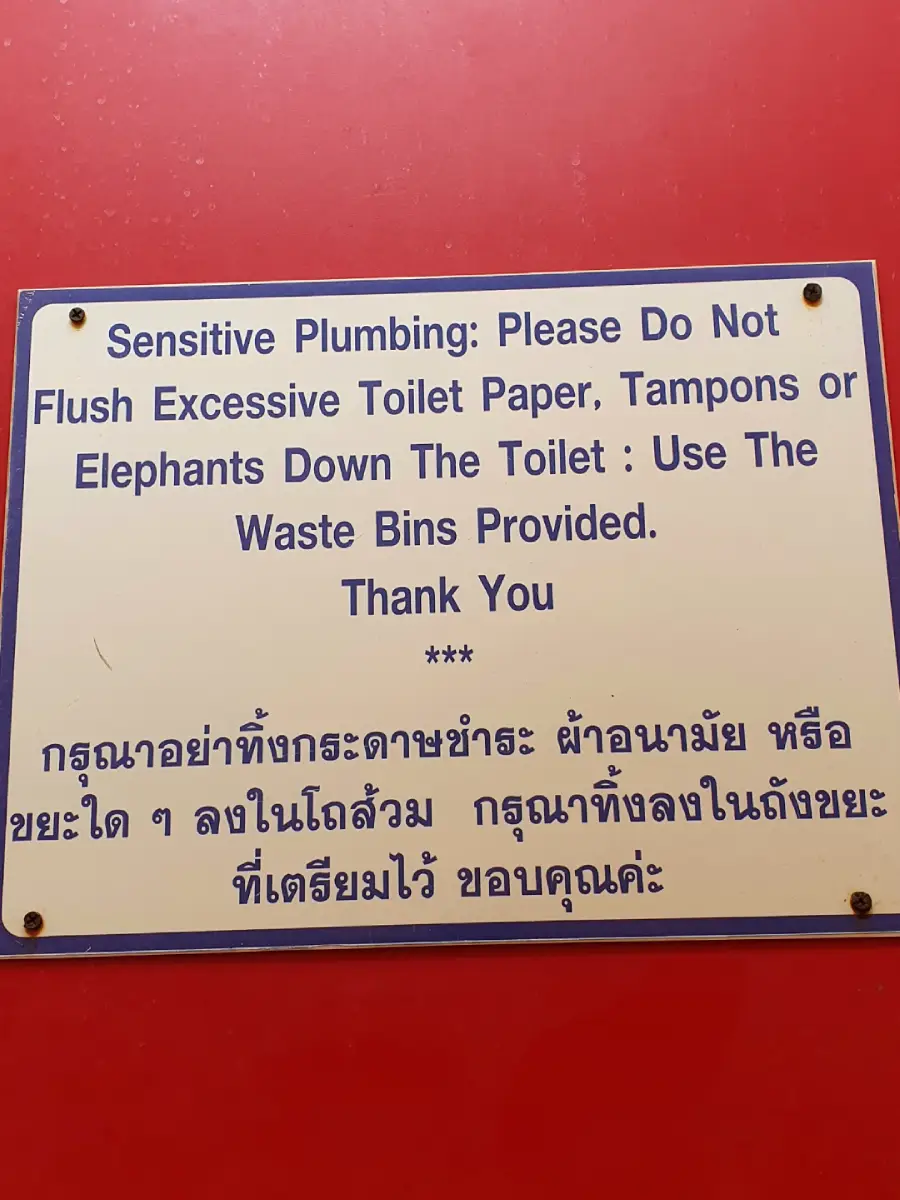 A sign on a red wall warns not to flush excessive toilet paper, tampons, or elephants, and asks to use waste bins instead. The message is written in both English and Thai.