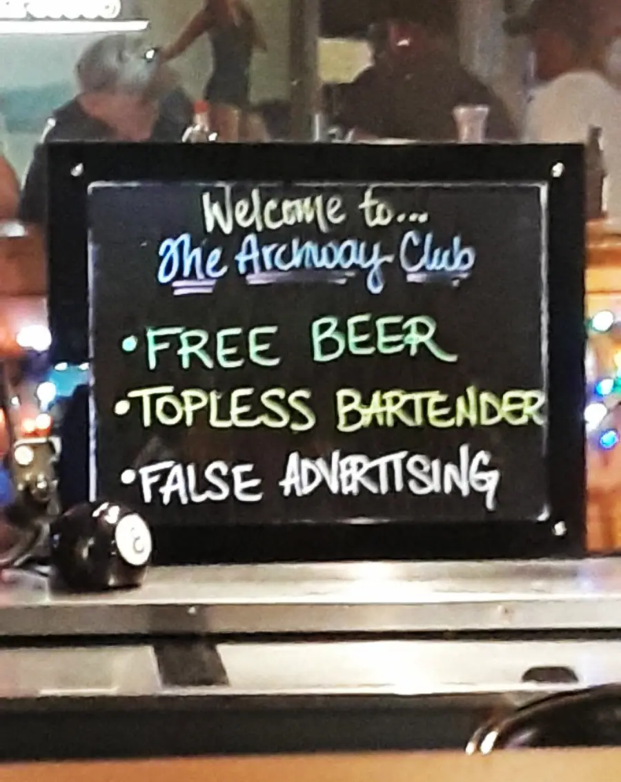 A chalkboard sign reads: “Welcome to The Archway Club. FREE BEER, TOPLESS BARTENDER, FALSE ADVERTISING,” with the text styled in different colors.