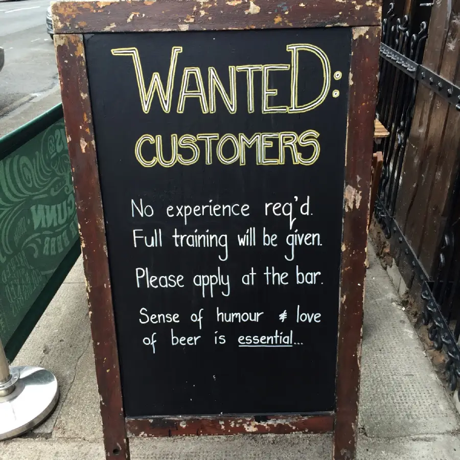 A chalkboard sign reads: “WANTED: CUSTOMERS. No experience req’d. Full training will be given. Please apply at the bar. Sense of humour & love of beer is essential...”