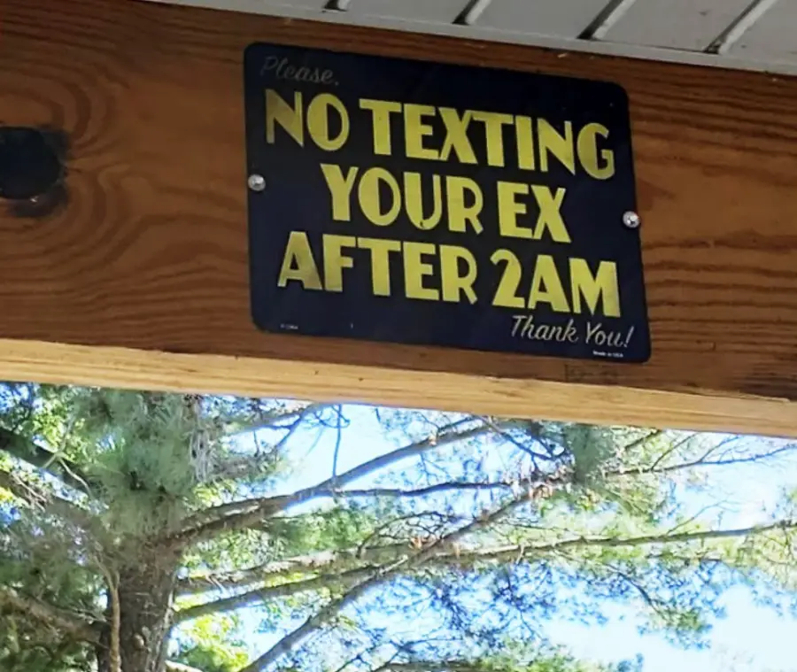A wooden sign reads, “Please. NO TEXTING YOUR EX AFTER 2AM. Thank you!” with trees and blue sky visible in the background.