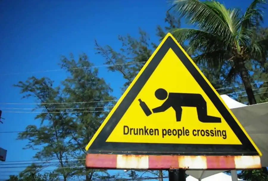 A yellow triangular sign shows a person crawling with a bottle, with text: “Drunken people crossing.” Palm trees and blue sky are visible in the background.