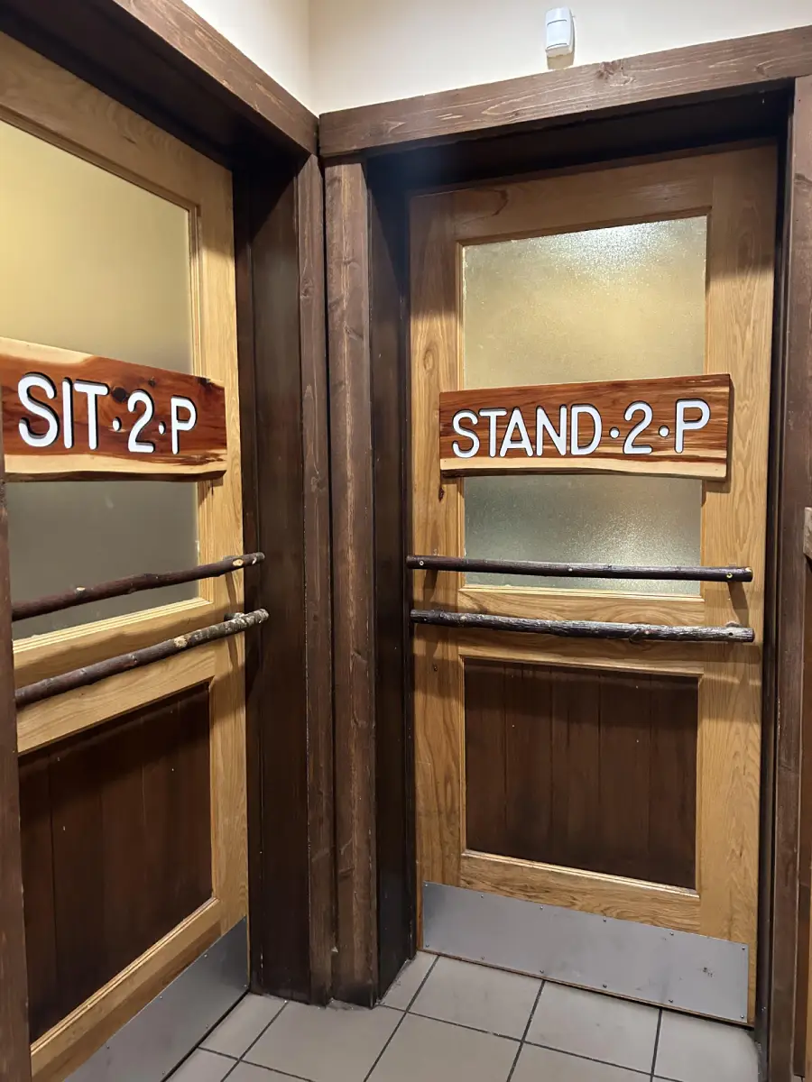 Two restroom doors: one labeled “SIT 2:P” and the other “STAND 2:P,” both with wooden signs and wooden rails, suggesting creative restroom gender signage.