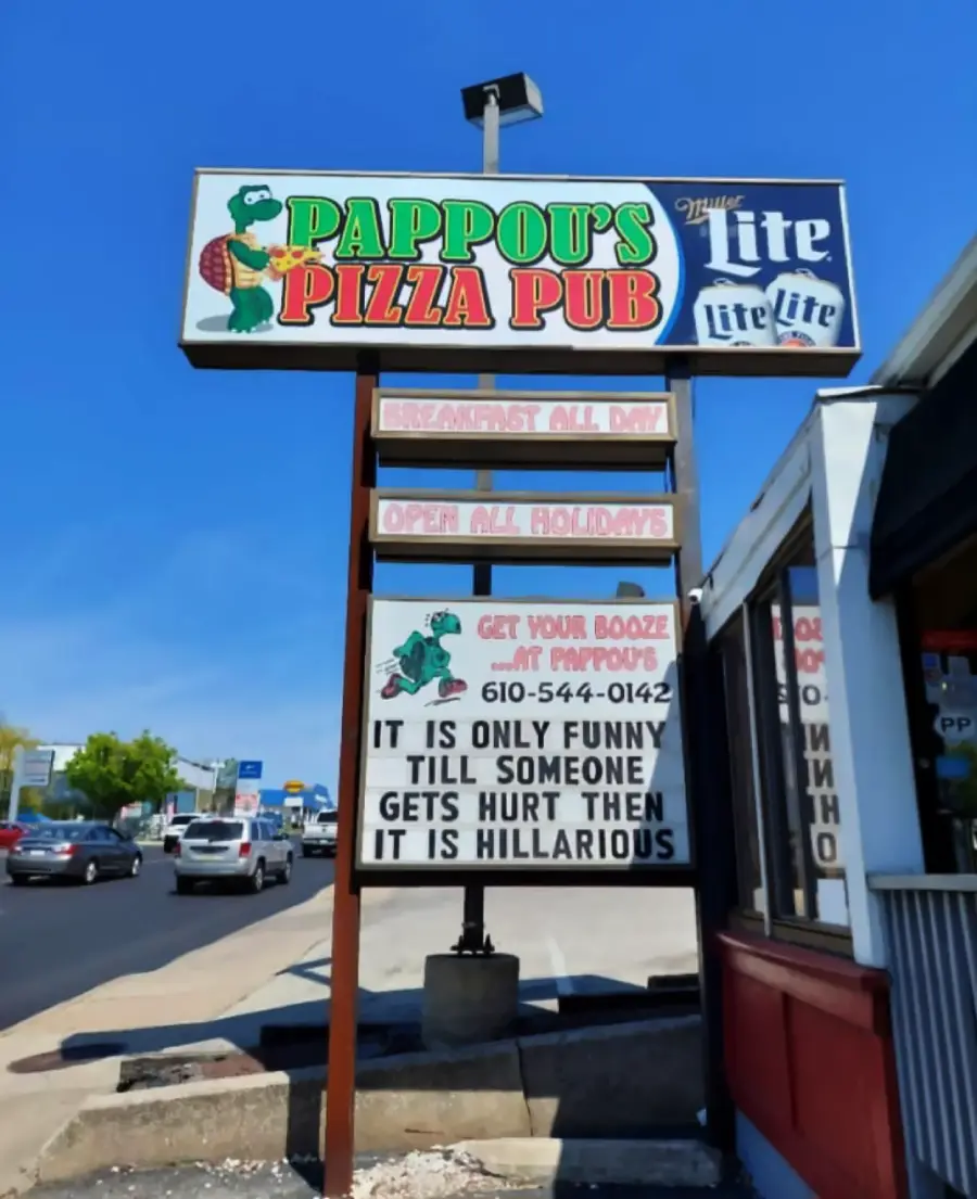 Pappou’s Pizza Pub sign with cartoon chef, beer ad, and message: “It is only funny till someone gets hurt then it is hillarious.” Street, cars, and blue sky in the background.