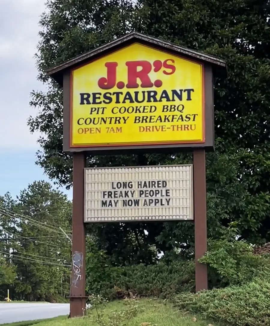 A roadside sign for J.R’s Restaurant reads: “LONG HAIRED FREAKY PEOPLE MAY NOW APPLY” below the main sign advertising pit cooked BBQ and country breakfast. Trees and power lines are in the background.