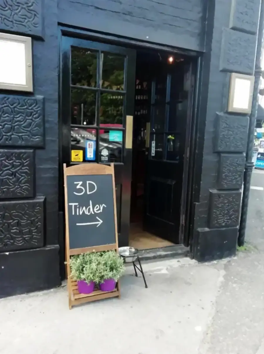 A black building with an open door displays a chalkboard sign reading “3D Tinder →” next to potted plants and a dog water bowl on the sidewalk.