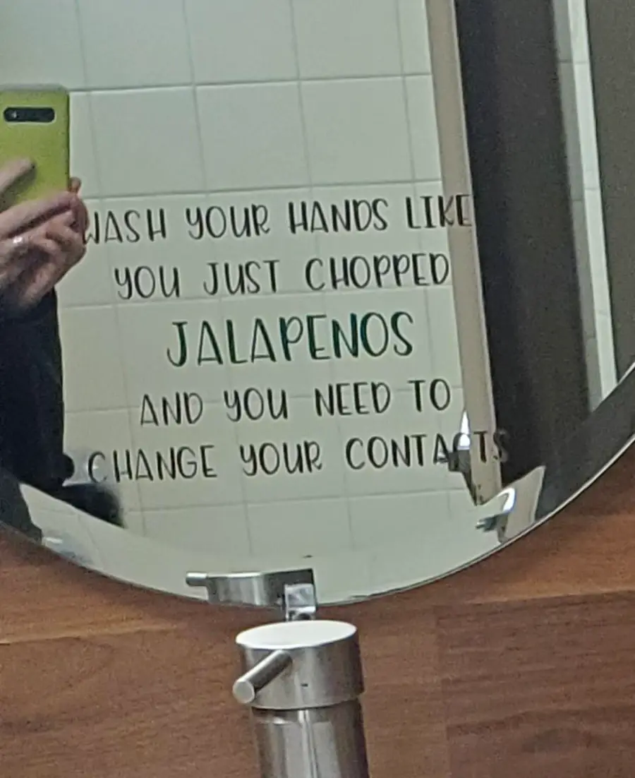 A mirror with text reading: “Wash your hands like you just chopped jalapenos and you need to change your contacts.” A person holding a green phone is partially reflected.