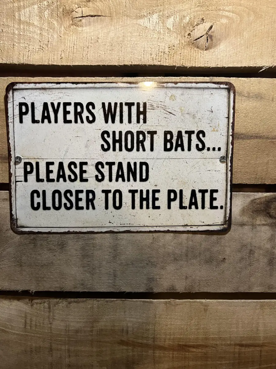 A metal sign on a wooden wall reads: “PLAYERS WITH SHORT BATS… PLEASE STAND CLOSER TO THE PLATE.”