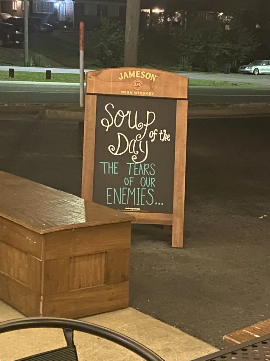 A chalkboard sign outside reads: “SOUP of the Day: THE TEARS OF OUR ENEMIES...” The sign is framed by a Jameson Irish Whiskey board on a street at night.