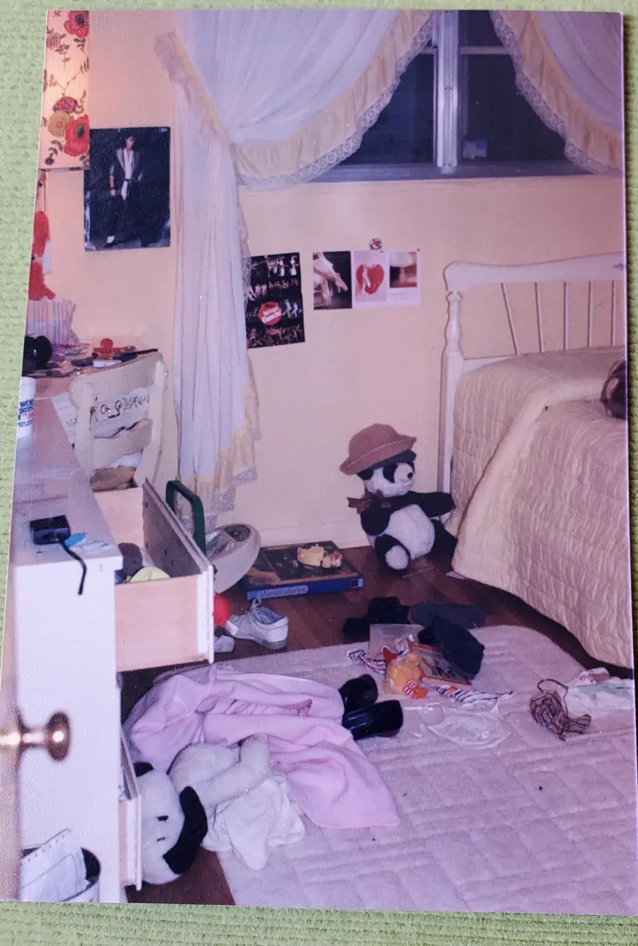 A messy bedroom with clothes and shoes scattered on the floor, a stuffed panda wearing a hat by the bed, posters on the wall, an open dresser drawer, and light curtains framing a window above the bed.