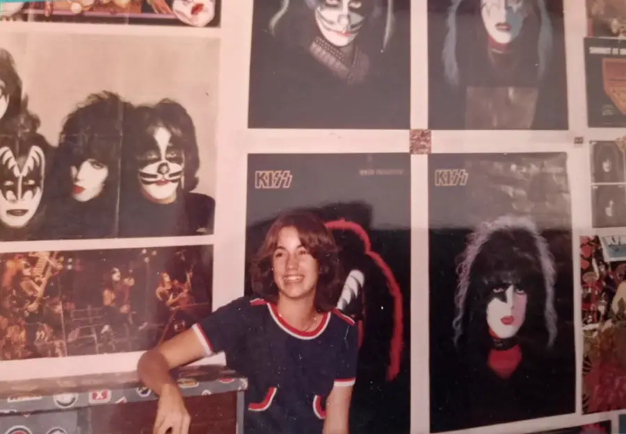 A smiling person with shoulder-length hair sits in front of a wall covered with posters of the band KISS, featuring the members in their signature makeup.