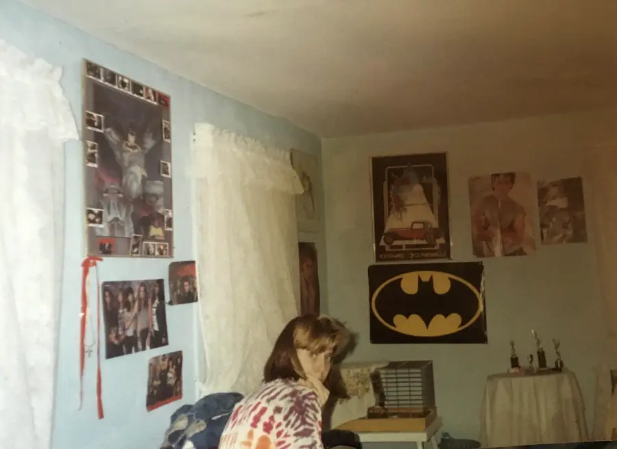 A person with long hair sits in a bedroom decorated with posters, including Batman and celebrities, white curtains on the windows, trophies on a round table, and a stack of books on a small cabinet.