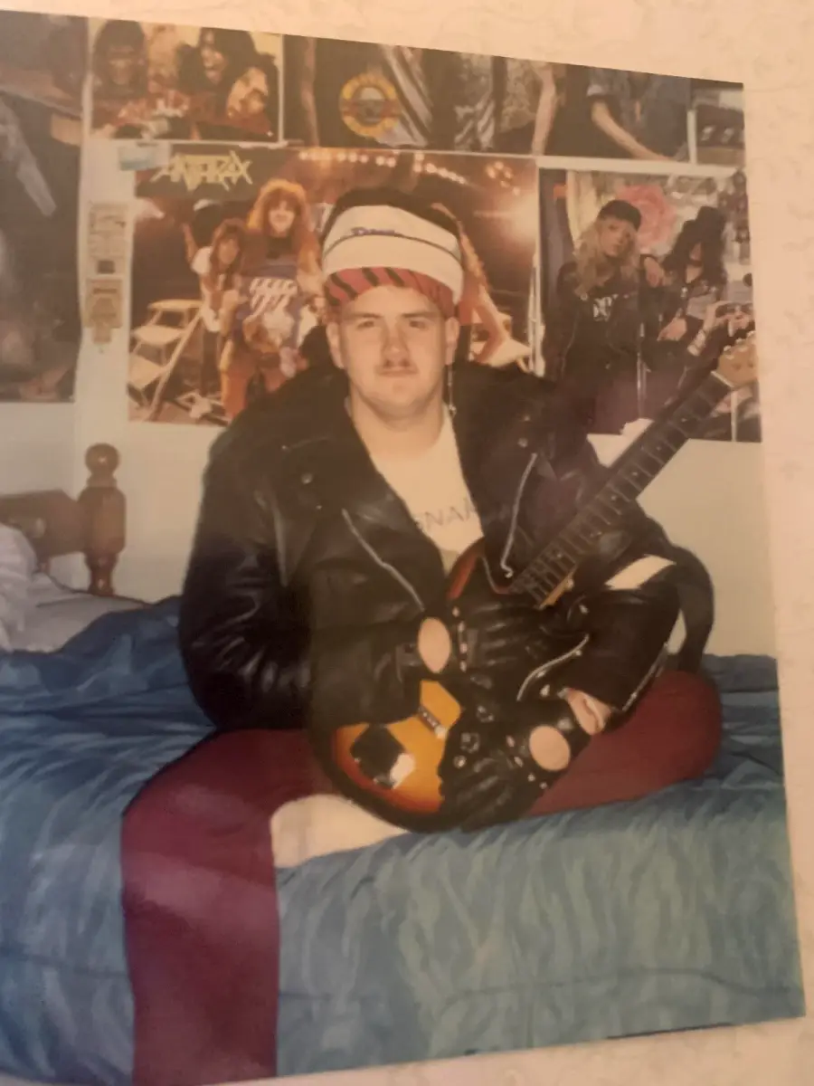 A person in a leather jacket and striped hat sits on a bed holding an electric guitar. Behind them, band posters cover the wall, creating a rock music-themed room.