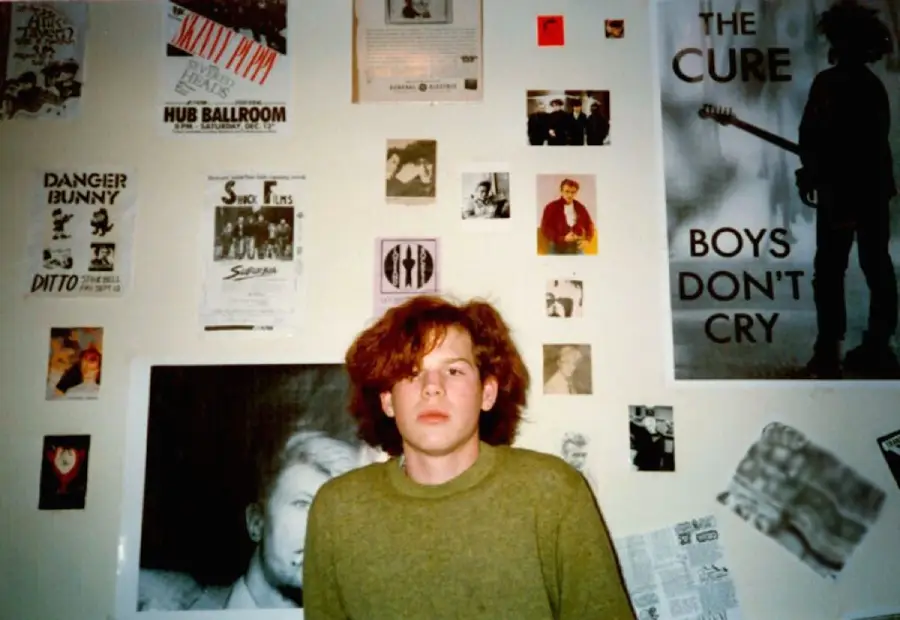 A young person with red hair in a green sweater sits in front of a wall covered with band posters, magazine clippings, and photos, including a large “The Cure: Boys Don’t Cry” poster.