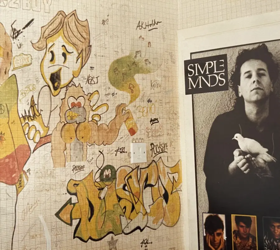 Wall with colorful graffiti and doodles, including a cartoonish face, abstract shapes, and layered tags. Beside it, a black and white Simple Minds band poster featuring a man holding a white dove.