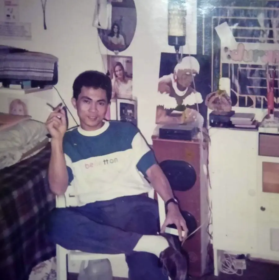 A man sits on a white chair indoors, holding a cigarette and smiling. The room has posters, a bed, speakers, a barred window, and various items on a table and shelf. The atmosphere feels casual and relaxed.