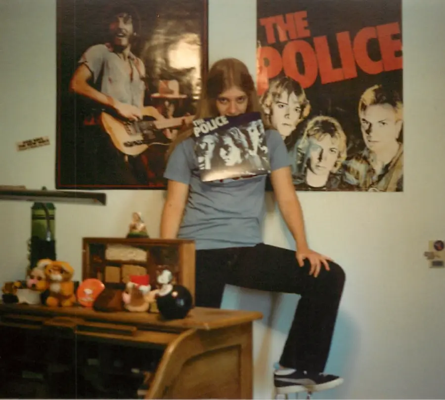 A person poses on a stool holding a “The Police” record in front of their face, with posters of musicians and The Police on the wall and plush toys on a shelf nearby.