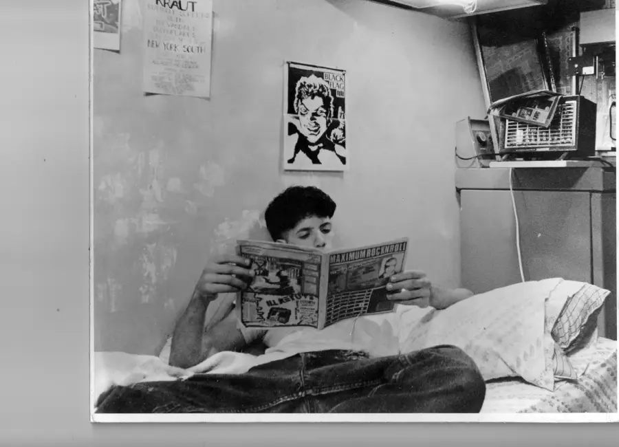 A person reclines on a bed reading a magazine. Posters and a radio are visible on the wall above. The room has a casual, lived-in feel, with pillows and a small shelf beside the bed.