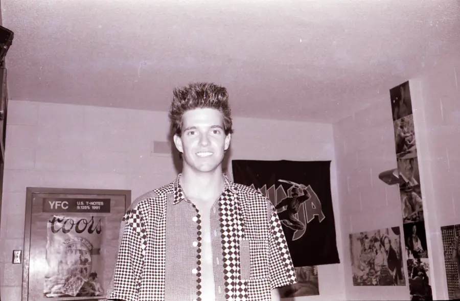 A young man with spiked hair, wearing a patterned shirt, smiles inside a room decorated with posters on the walls. The photo is in black and white.