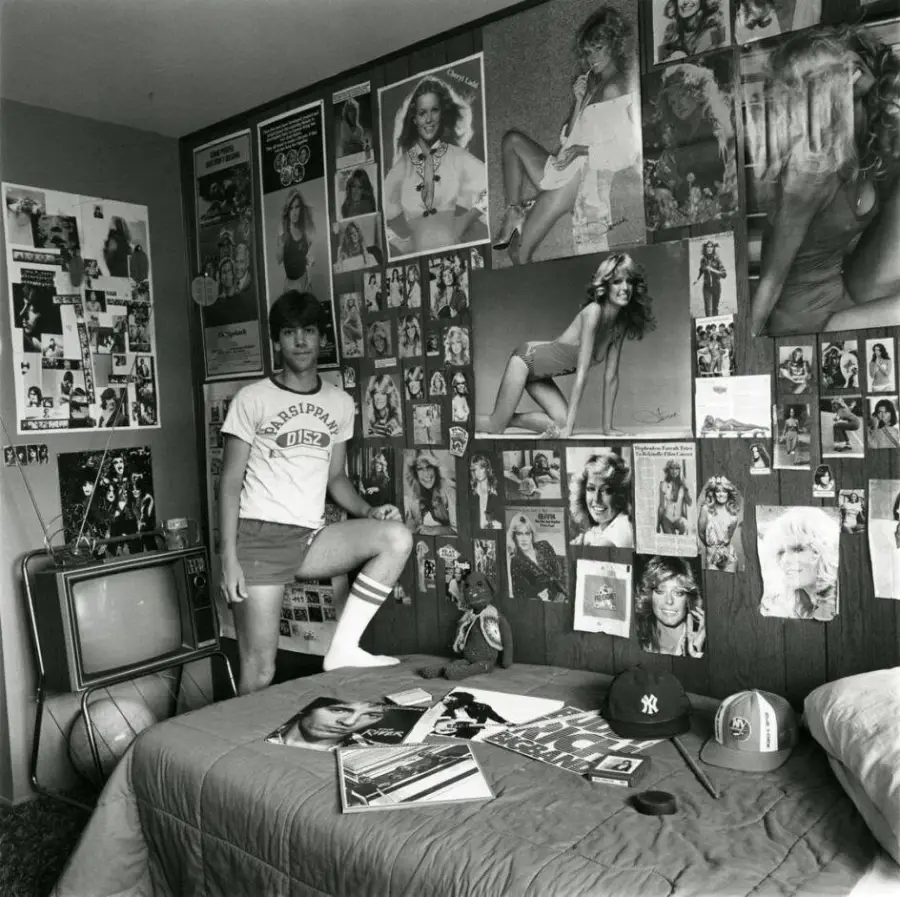 A teen stands in a bedroom with walls covered in posters of women. The room has a bed, magazines, a TV, and baseball caps. The scene appears to be from the 1970s or 1980s.