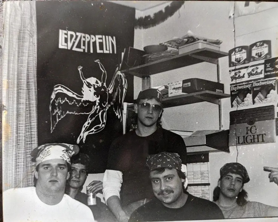 Five men in bandanas sit in a small room with shelves, a Led Zeppelin poster, and beer boxes on the wall. The scene is casual and appears to be from the 1980s or 90s.