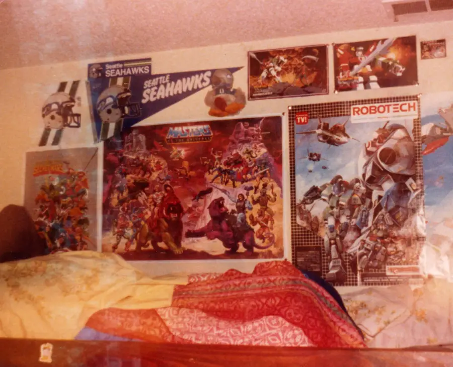 A bedroom wall filled with 1980s posters, including Seattle Seahawks, Transformers, Masters of the Universe, Robotech, and more. A colorful bedspread covers a bed in the foreground.
