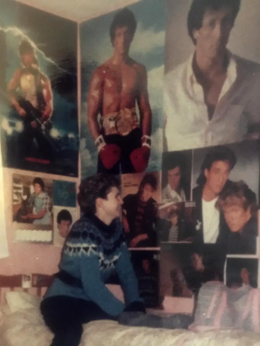 A person sits on a bed in a room decorated with posters of a male actor, mostly in boxing and dramatic poses, covering the walls. The person wears a patterned sweater and looks toward the posters.