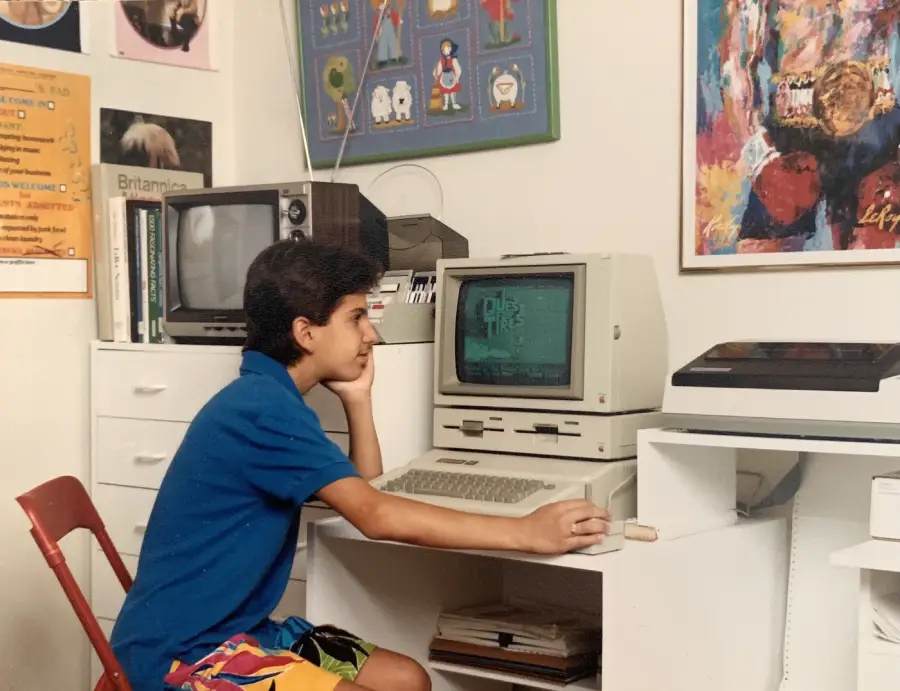 A boy in colorful shorts and a blue shirt sits at a desk using an old beige computer, with a monitor, keyboard, and joystick. The room has posters, a TV, and colorful artwork on the walls.