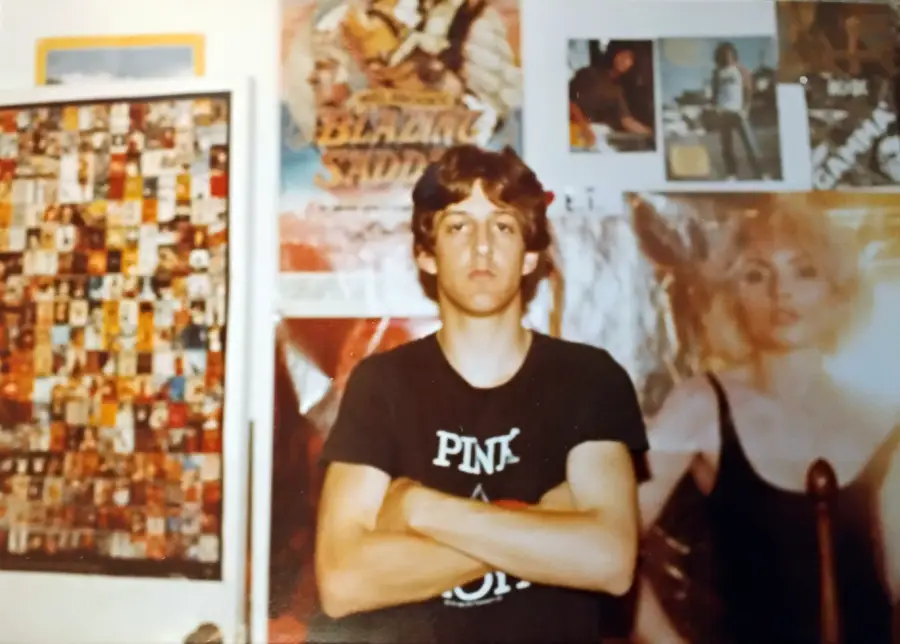 A young person with arms crossed stands in front of a wall covered with posters and photos, including a large image of a woman in a black top. The person wears a dark “Pink Floyd” T-shirt and has short, light-brown hair.