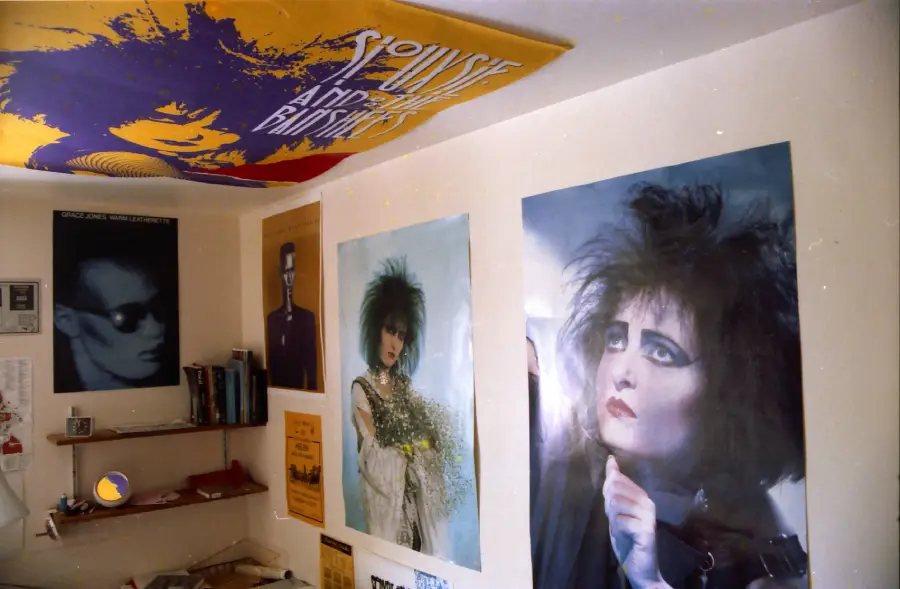 A room with posters of goth musicians, including Siouxsie Sioux, on the walls and ceiling. A small shelf with books and a mirror are visible, creating a distinctive alternative aesthetic.