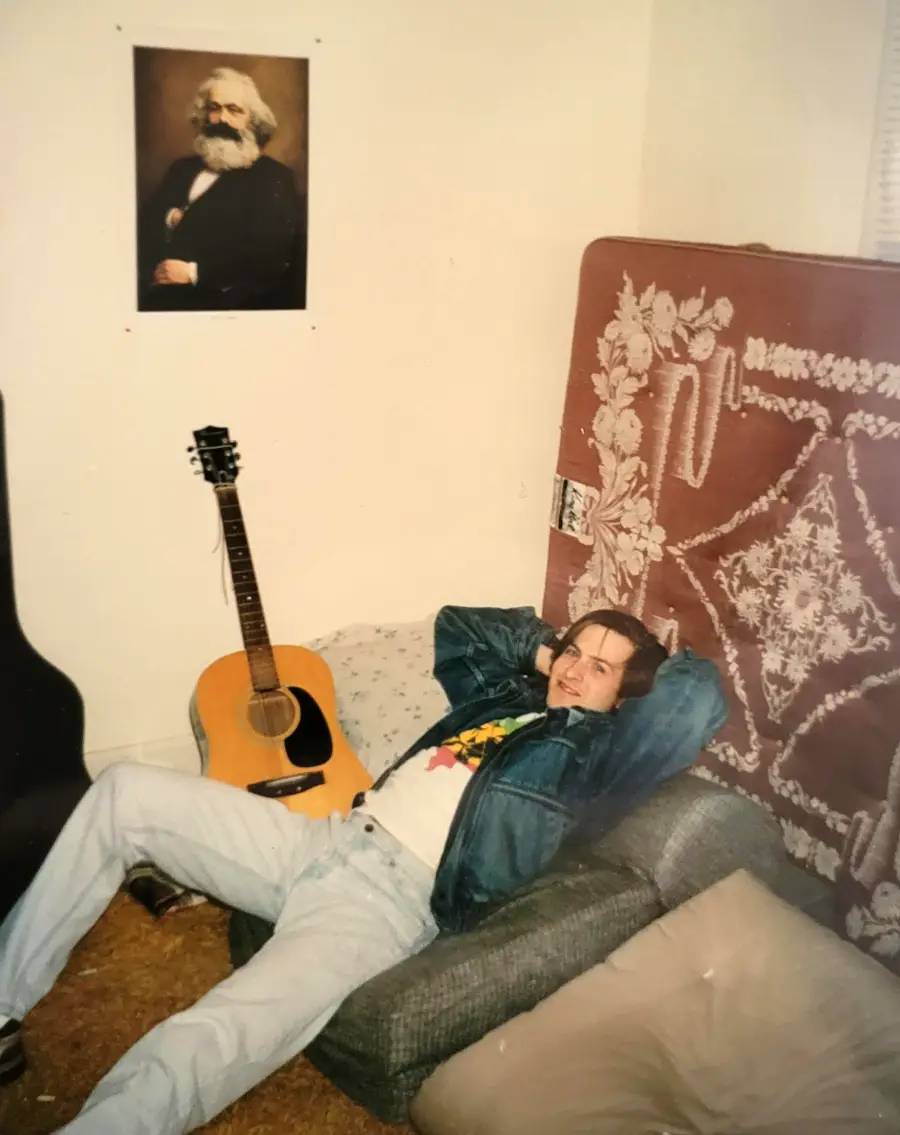 A person lounges on a sofa in a casual room with a guitar, a mattress with a patterned blanket, and a wall poster of an older man with a beard.