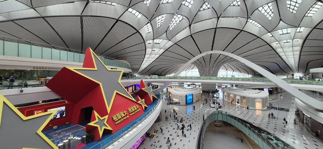 Modern airport interior with a unique arched ceiling, large red and yellow star decorations, people walking, shops, and glass railings. The architecture is spacious and futuristic.