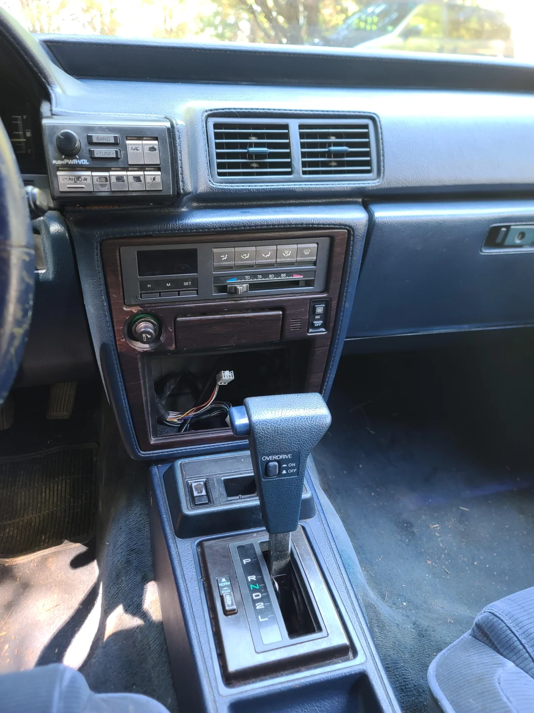 Close-up of a vintage car’s center console showing an automatic gear shift, classic radio, and climate controls. The dashboard and trim are dark blue with wood accents, and sunlight filters in from the window above.