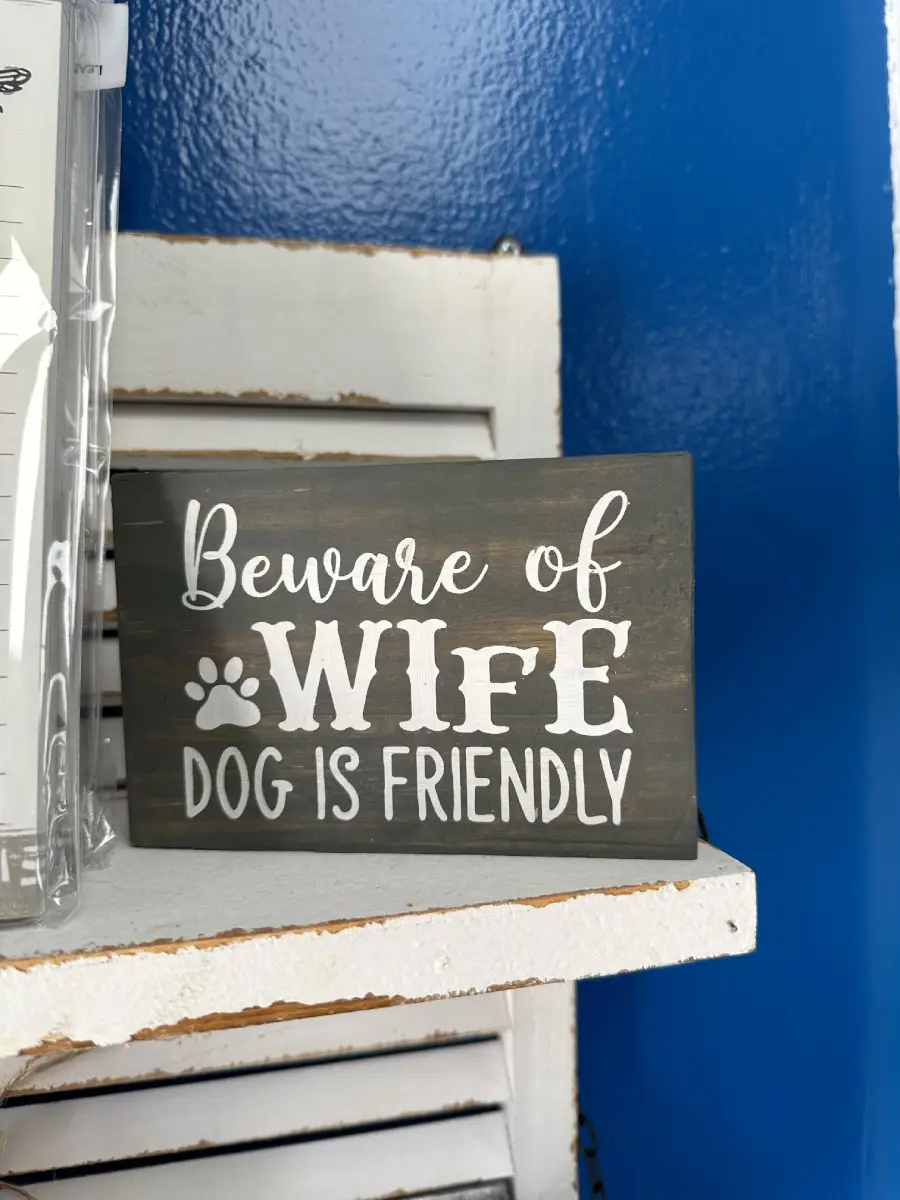 A sign reads “Beware of Wife, Dog is Friendly,” with a paw print graphic; it sits on a white shelf against a blue wall.