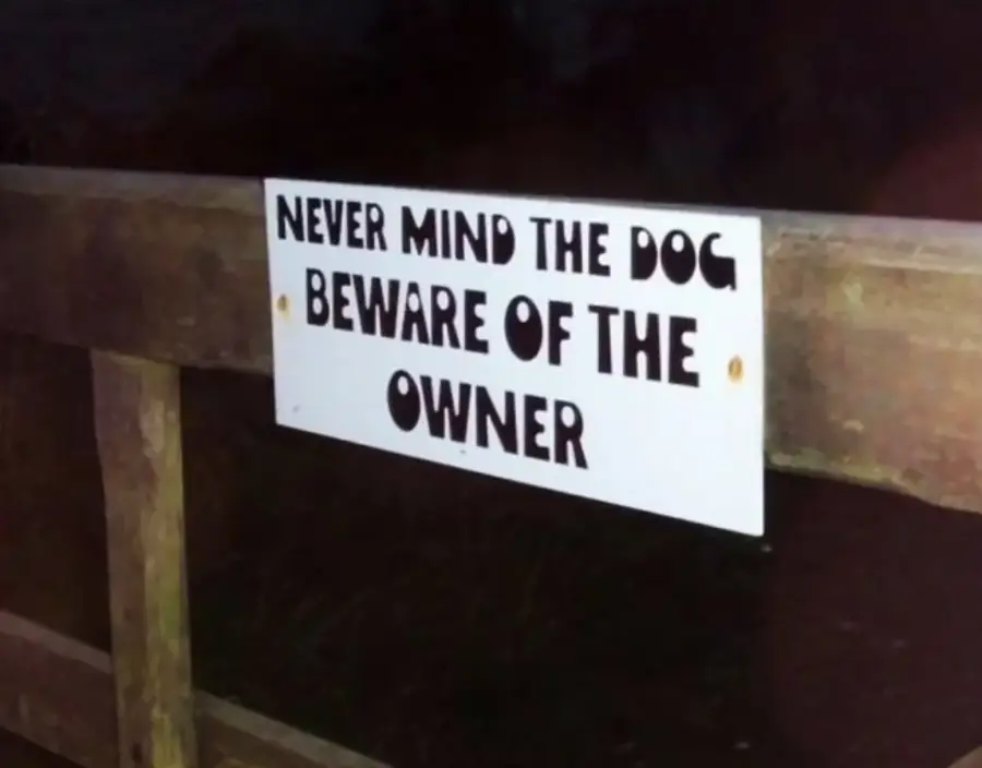 A sign on a wooden fence reads, “Never mind the dog, beware of the owner,” in bold black letters on a white background.