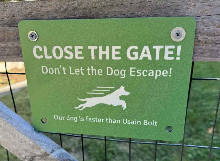 A green sign on a gate reads, “CLOSE THE GATE! Don’t Let the Dog Escape! Our dog is faster than Usain Bolt,” with an illustration of a running dog.