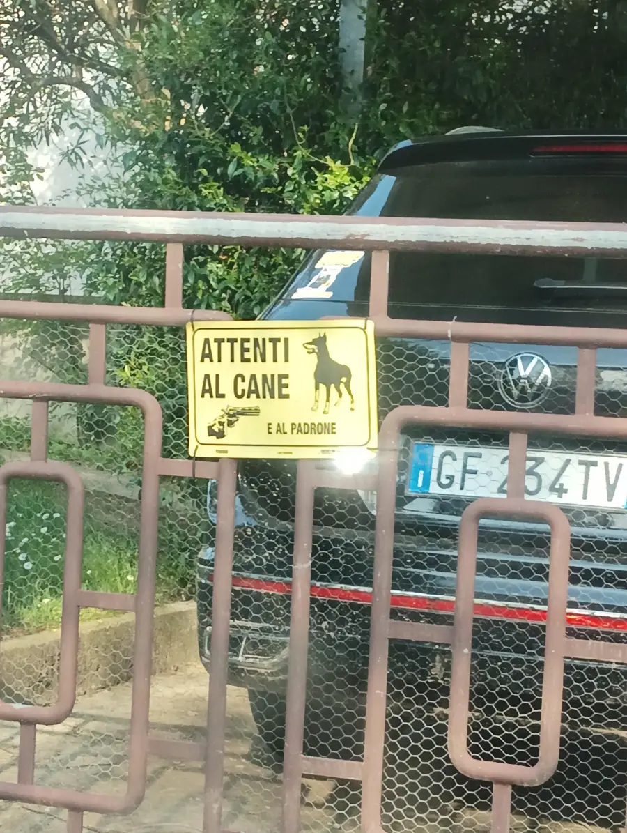 A yellow sign on a gate reads “Attenti al cane e al padrone” with an image of a dog and man, warning about the dog and its owner. A black car with a visible partial license plate is parked behind the gate.