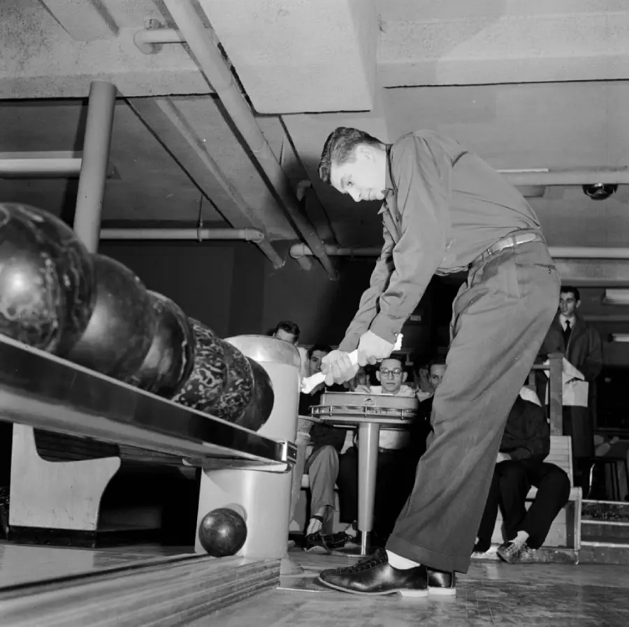 A man prepares to bowl in a bowling alley, leaning forward to pick up a ball as several people watch from the seating area behind him. Black and white photo.