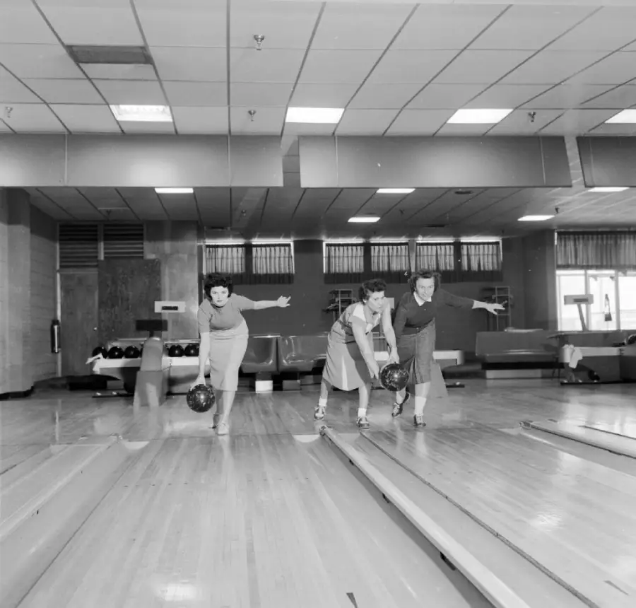 Three women are bowling side by side in a bowling alley, each holding a ball and mid-swing. The scene is in black and white, showing lanes, pins, and seating in the background.