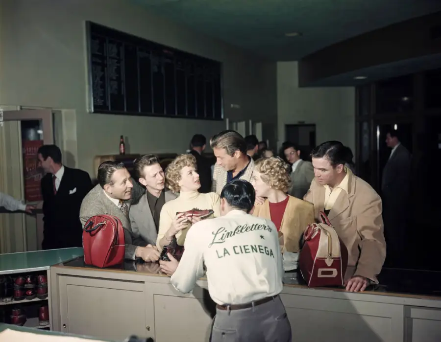 A group of people chat and smile at a counter, with a person in a “Linkletter’s La Cienega” jacket serving them food or drinks in a lively indoor setting.