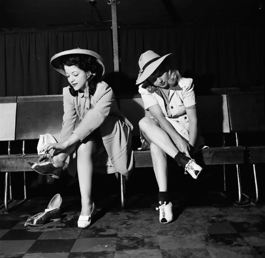 Two women sit on a bench, wearing dresses and hats, as they change into roller skates. The background is dark, and they appear to be in a roller rink or similar indoor venue.