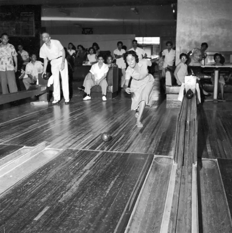 A woman bowls on a wooden lane as people watch from behind. A man lines up to bowl as well. The setting appears to be a vintage, crowded bowling alley.