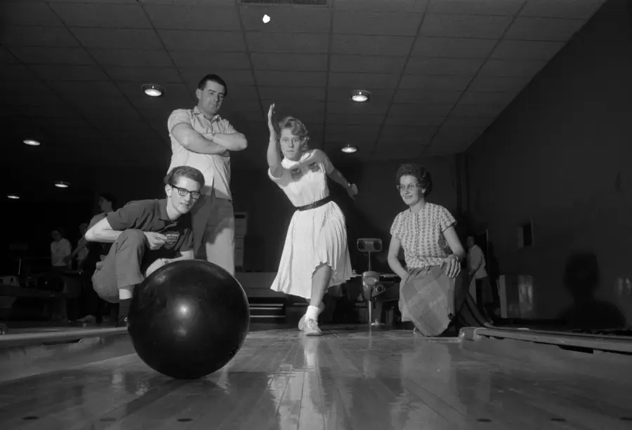 A woman in a dress bowls while three people watch her closely. The bowling ball rolls down the lane toward the pins in a dimly lit bowling alley.
