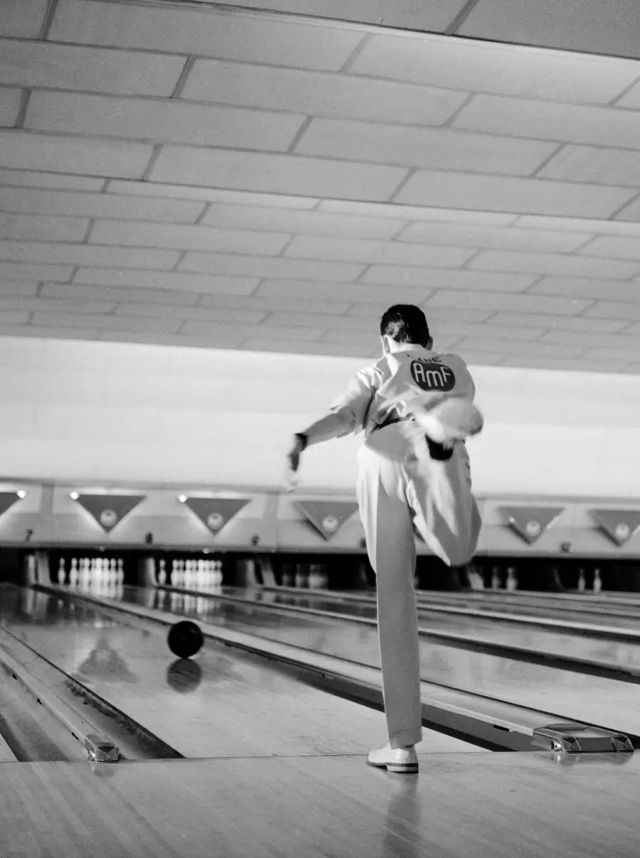 A person in a bowling alley, wearing a jacket with “AMF” on the back, releases a bowling ball down the lane, standing on one leg with the other leg raised behind them.