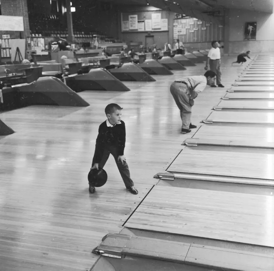 A young boy prepares to bowl at a vintage bowling alley, while other people bowl in adjacent lanes. The setting features wooden floors and a classic atmosphere.