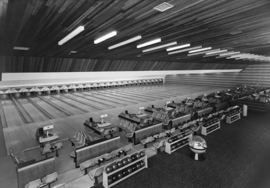Black and white photo of a retro bowling alley with multiple empty lanes, ball returns, and seating areas. The wooden ceiling and vintage furniture add to the classic atmosphere.