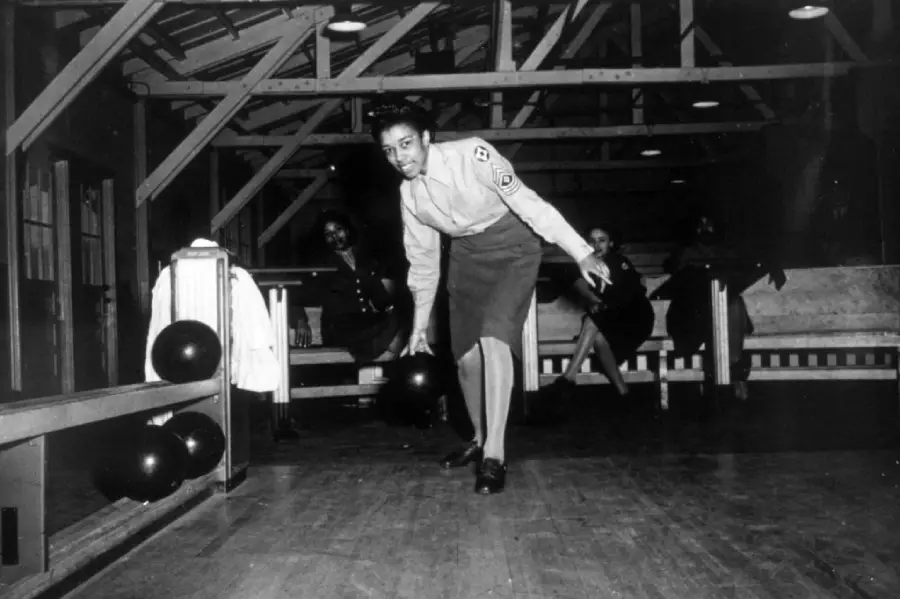 A woman in a military uniform bowls on a wooden lane indoors, while two others sit and watch in the background under exposed beams. Bowling balls are visible on the side.
