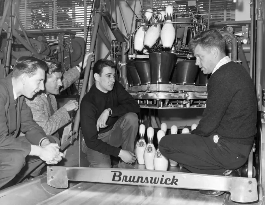 Four men examine a bowling pin setting machine marked “Brunswick.” Two men crouch, one holds a pin, while another man sits and one kneels. Bowling pins and machinery are visible in the background.