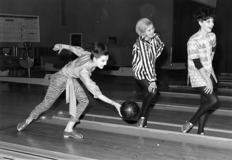 Three women at a bowling alley, with one woman in striped pants releasing a bowling ball, while the other two, dressed stylishly, watch and smile. The scene appears lively and fun.